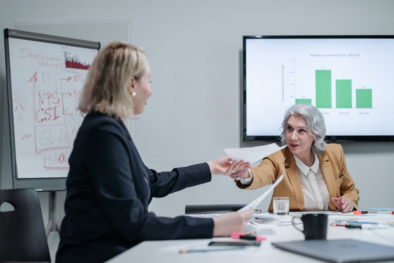 Duas mulheres, uma empresária idosa e uma mais nova, em uma mesa de reunião com gráficos ao fundo, representando a passagem de bastão na sucessão familiar de uma empresa.