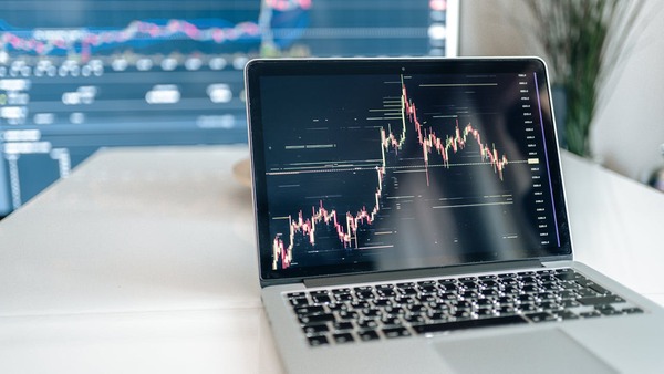 Notebook sobre mesa de escritório exibindo gráfico de oscilação de dados com telão azul de indicadores desfocado ao fundo.