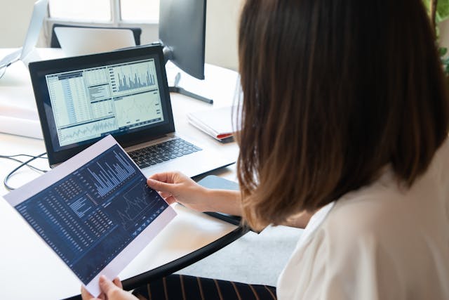 Profissional de costas analisando dados, relatórios e gráficos financeiros no notebook e em folha de papel para planejamento orçamentário.
