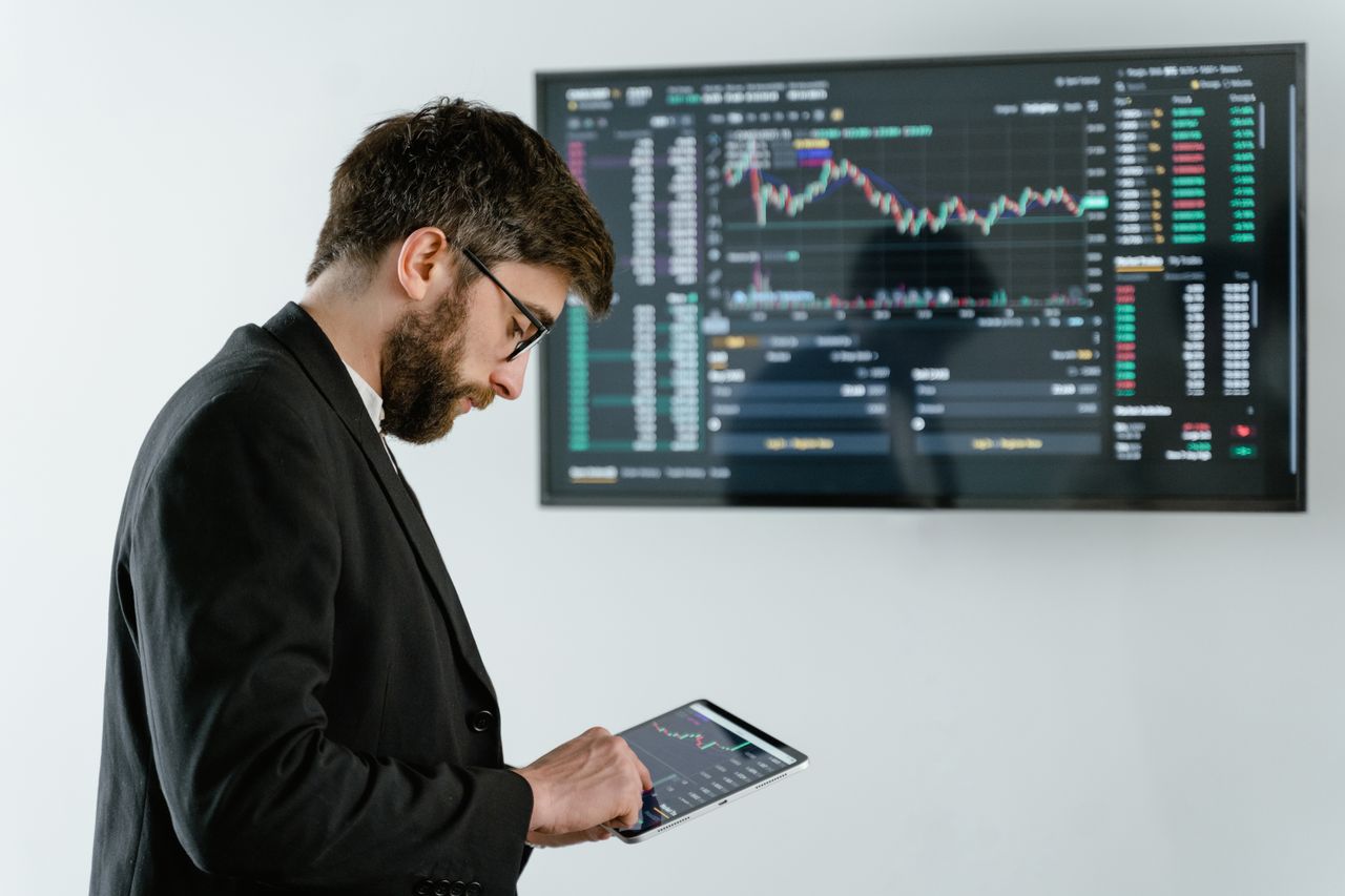 Executivo jovem e sério analisa dados financeiros em um tablet, com tela de fundo exibindo gráficos estatísticos de faturamento e custos corporativos.