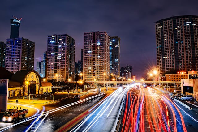 Fotografia noturna em longa exposição da Avenida Paulista com linhas de luz coloridas em movimento e prédios comerciais iluminados ao fundo.